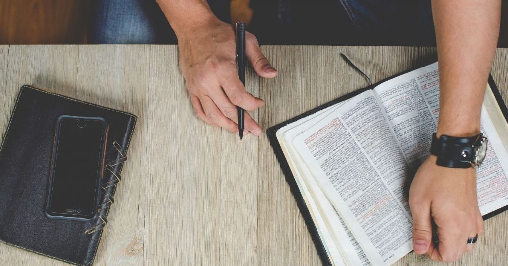 photo of a man reading a bible at a table with pen